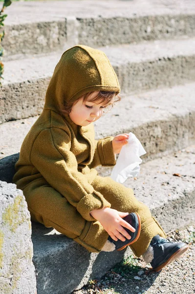 かわいい幼児の子供は ぬれたワイプで靴を掃除し 公共公園の階段に座って 緑の暖かいジャンプスーツを着て — ストック写真