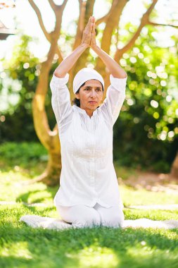 50-55 yaşında olgun bir kadın, yeşil güneşli bahçede meditasyon yapıyor, sağlıklı bir yaşam tarzı var.