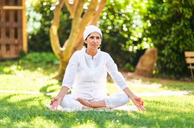 50-55 yaşında olgun bir kadın, yeşil güneşli bahçede meditasyon yapıyor, sağlıklı bir yaşam tarzı var.