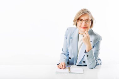 Beyaz arka planda poz veren orta yaşlı bir kadının stüdyo portresi, masada oturuyor ve notlar alıyor.