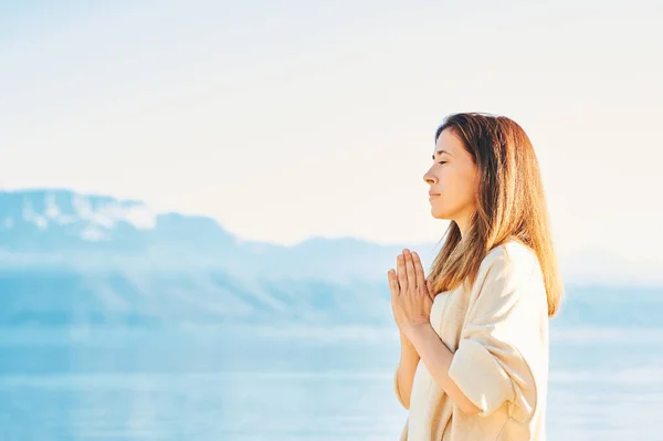 Beautiful spiritual woman meditating by the lake, wearing beige clothes