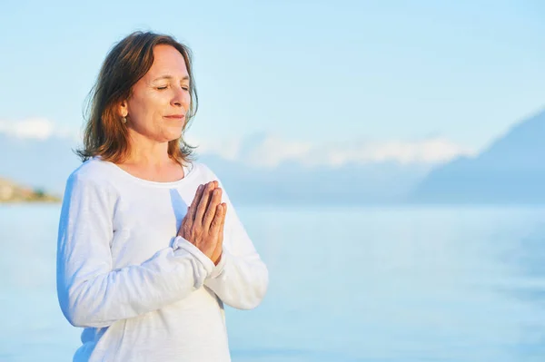 Beautiful spiritual woman meditating by the lake, wearing white clothes