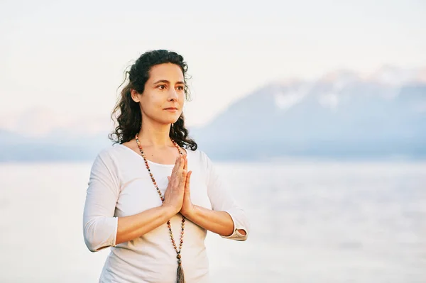 Beautiful spiritual woman meditating by the lake, wearing white clothes