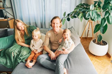 Dört kişilik mutlu bir aile kanepede dinleniyor, çift küçük kız ve bebekle oynuyor.