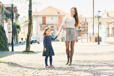 Outdoor portrait of happy young mother and little daughter walking down the street, early spring or fall weather