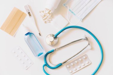 Tıbbi geçmiş, stetoskop, dijital termometre, yüz maskesi ve beyaz arka planda haplar.