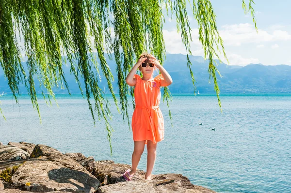 Turuncu elbiseli, güzel güneşli bir günde gölün yanında duran sevimli küçük bir kızın yaz portresi. — Stok fotoğraf