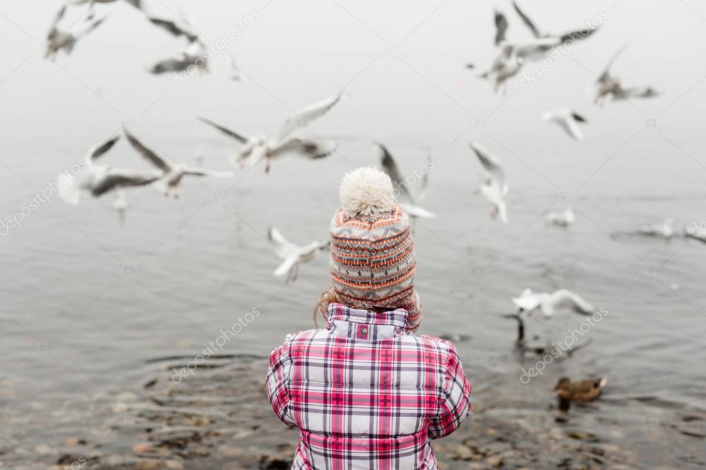 Little girl feeding birds on the lake in winter time, wearing warm jacket and hat