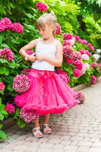 Güzel küçük kız parlak pembe etek ve beyaz bluz giyiyor, ortanca çalılarının yanında dikey portre — Stok fotoğraf