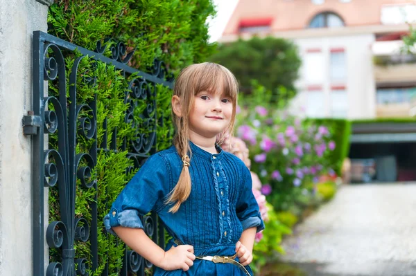 Portrait extérieur d'une belle petite fille portant une robe en jean, debout à côté de la porte, avec maison sur fond — Photo