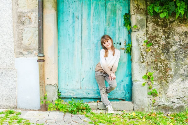 Außenporträt eines niedlichen kleinen Mädchens, das neben der türkisfarbenen Tür steht, beigen Pullover, Hose und Schuhe trägt — Stockfoto