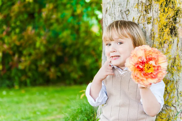 Outdoor portrait of a cute toddler boy on a nice sunny day, sitting under the tree, holding beautiful peony flower — Stock Photo, Image