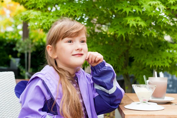 Открытый портрет дошкольницы в дождевом пальто, сидящей в кафе с мороженым и горячим шоколадом на столе — стоковое фото