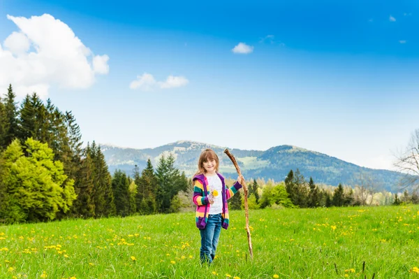 Cute little girl walking in mountains, holding wooden stick and wildflowers, wearing jeans and colorful jacket — Stock Photo, Image