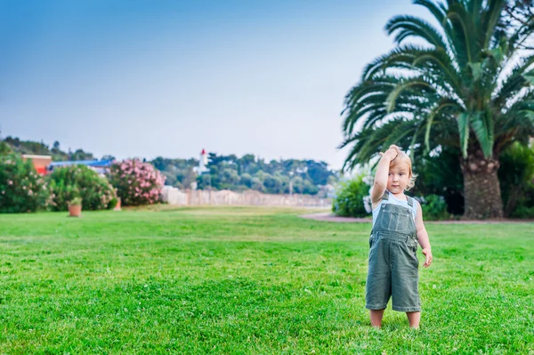 Söta barn pojke stående på en gräsmatta, Palm och stranden på bakgrund, iklädd overall — Stockfoto