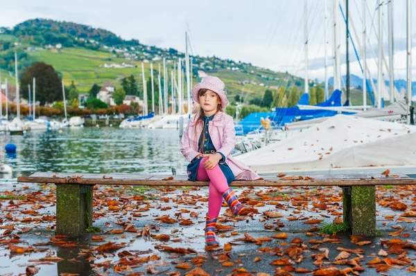 Schattig klein meisje zittend op een bankje op een mooie herfstdag, met roze vinyl jas en muts op — Stockfoto