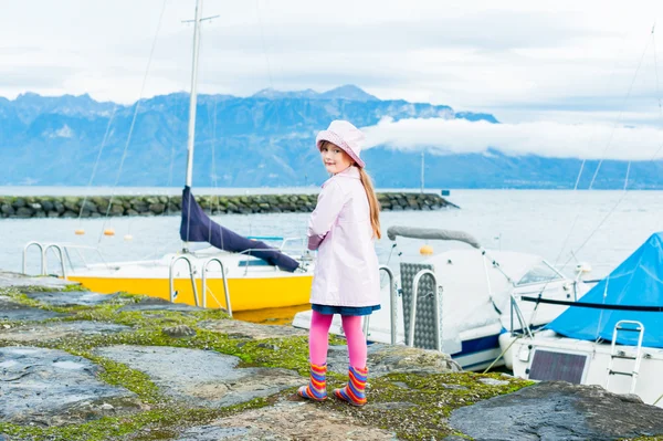 Venkovní portrét roztomilá holčička, nosí růžové vinyl dešti kabát a klobouk, barevný déšť boty a růžové punčocháče — Stock fotografie