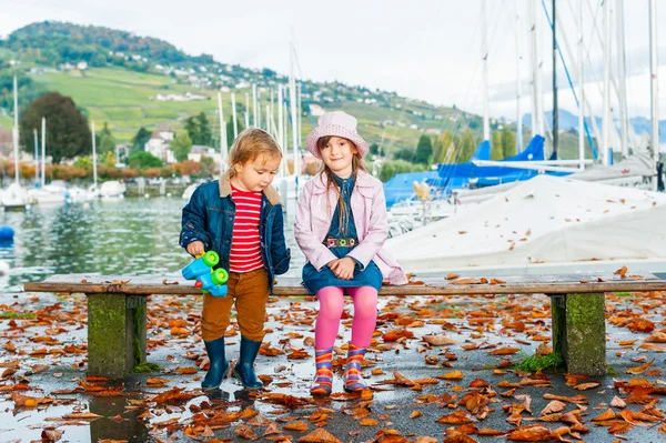 Hermanita y hermano pequeño jugando juntos al lado del lago, sentados en un banco en un cálido día de otoño — Foto de Stock