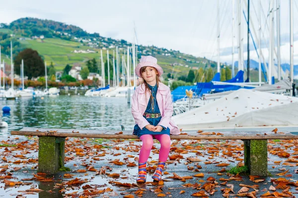 Schattig klein meisje zittend op een bankje op een mooie herfstdag, met roze vinyl jas en muts op — Stockfoto