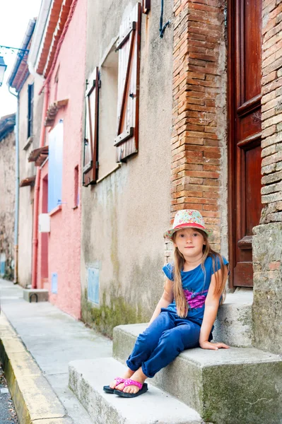 Красивая маленькая девочка сидит на ступеньке, в синих брюках и топе, шляпа с цветами — стоковое фото