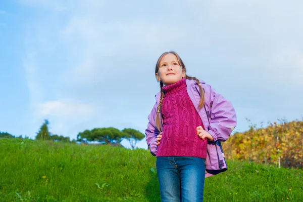 Autumn portrait of a cute little girl in pink pullover, jeans and violet rain jacket, green grass and blue sky on background — Stock Photo, Image