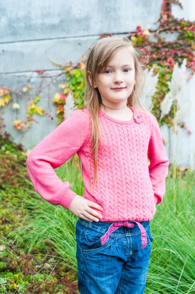 Außenporträt eines niedlichen kleinen Mädchens in einem Garten an einem schönen Herbsttag, mit grauem Mantel, Jeans, rosa Pullover — Stockfoto