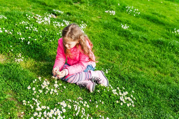 Menina bonita brincando com flores em um parque em um dia ensolarado agradável em um início de primavera, vestindo jaqueta rosa brilhante — Fotografia de Stock