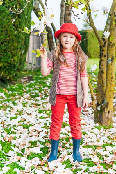 Ritratto primaverile di una graziosa bambina con cappello rosso, jeans e giacca a maglia, magnolia sullo sfondo — Foto Stock