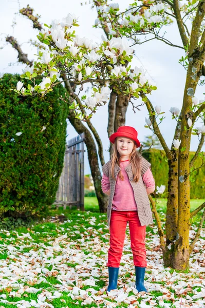 Frühlingsporträt eines süßen kleinen Mädchens mit rotem Hut, Jeans und Strickjacke, Magnolie auf dem Hintergrund — Stockfoto