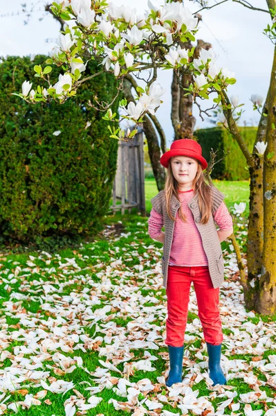 Spring portrait of a cute little girl in a red hat, jeans and knitted jacket, magnolia on background — Stock Photo, Image