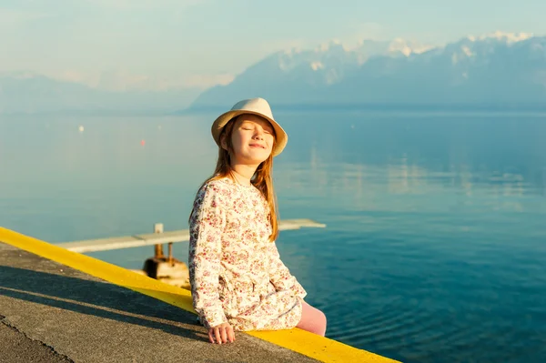 Retrato al atardecer de una adorable niña sentada junto al lago con los ojos cerrados y sonrisa en la cara, con sombrero y hermoso vestido — Foto de Stock