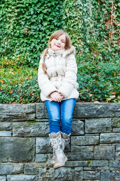 Retrato al aire libre de una linda niña sentada en una pared, con chaqueta blanca cálida, jeans y botas —  Fotos de Stock