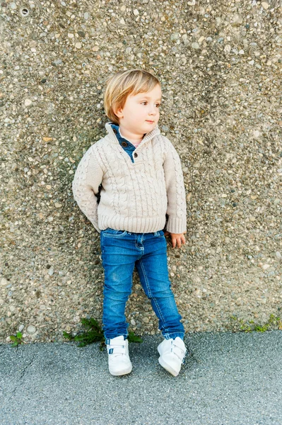 Retrato al aire libre de un lindo niño de pie junto a la pared de piedra, usando jersey beige y jeans — Foto de Stock