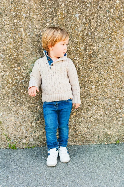Outdoor portrait of a cute toddler boy standing next to stone wall, wearing beige pullover and jeans — Stock Photo, Image