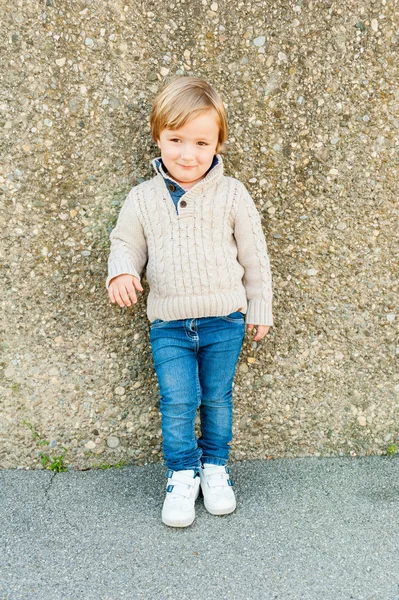 可爱的蹒跚学步的男孩站在旁边的石头墙，穿着米色套衫和牛仔裤的室外肖像 — 图库照片