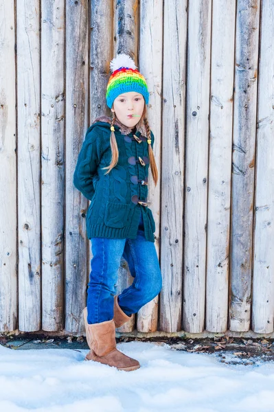 Vinter porträtt av en söt liten flicka, står bredvid trävägg, bär, grön kofta, jeans, bruna stövlar och färgglada hatt — Stockfoto
