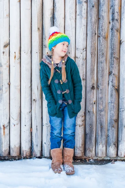 Vinter porträtt av en söt liten flicka, står bredvid trävägg, bär, grön kofta, jeans, bruna stövlar och färgglada hatt — Stockfoto