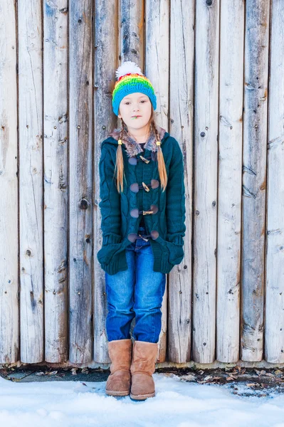 Vinter porträtt av en söt liten flicka, står bredvid trävägg, bär, grön kofta, jeans, bruna stövlar och färgglada hatt — Stockfoto