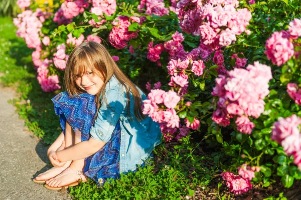 Portret van een schattig klein meisje op zonsondergang, zittend naast prachtige rozenstruiken — Stockfoto