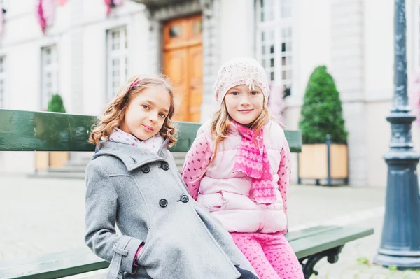 Outdoor Portret van twee schattige kleine meisjes van zeven jaar oud, rust op de Bank in een stad — Stockfoto