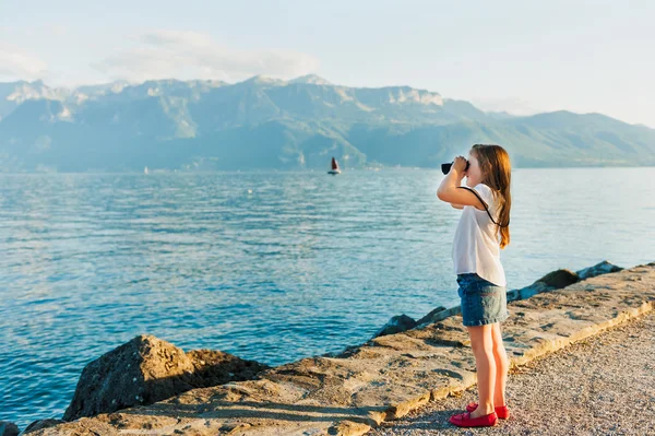 Cute little girl standing next to beautiful lake on sunset and looking in binoculars — Stock Photo, Image