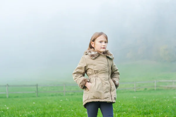 Außenporträt eines niedlichen kleinen Mädchens in einem Garten an einem nebligen Tag, das einen warmen beigen Mantel trägt — Stockfoto