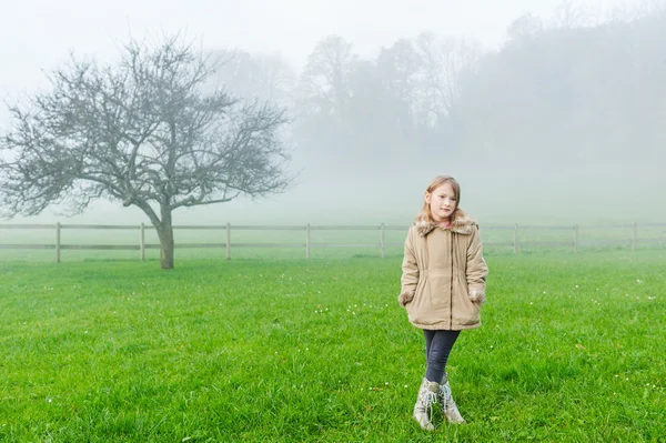 Utomhus porträtt av en söt liten flicka i en trädgård på en dimmig dag, klädd i varma beige kappa — Stockfoto