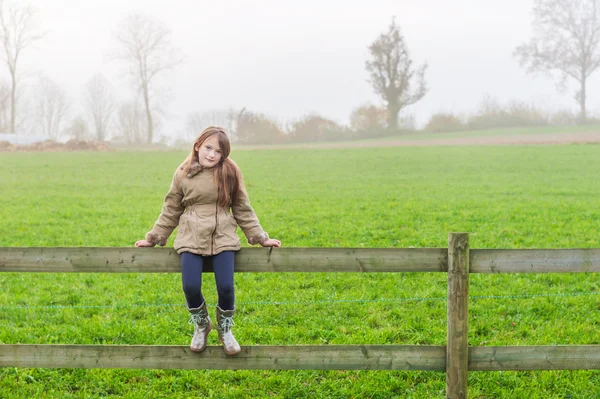 Niedliches kleines Mädchen sitzt auf einem Zaun in einer Landschaft an einem kalten nebligen Tag — Stockfoto