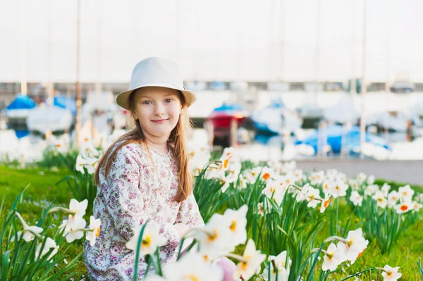 Spring portrait of adorable little girl by the lake, resting in a park between white narcissus, wearing a hat, sunset — Stock Photo, Image