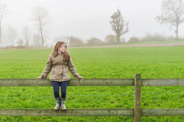 Söt liten flicka sitter på ett staket i en landsbygd på en kall dimmig dag — Stockfoto