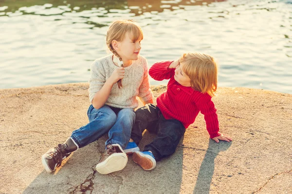 Duas crianças, menina e menino descansando no lago ao pôr-do-sol, usando pulôveres e jeans, imagem tonificada — Fotografia de Stock