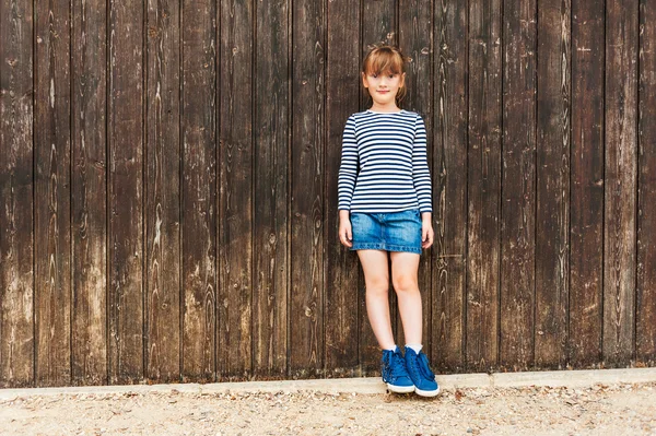 Ritratto all'aperto di una bambina carina, vestita con abito, gonna in denim e belle scarpe da ginnastica blu, in piedi contro il muro di legno marrone — Foto Stock