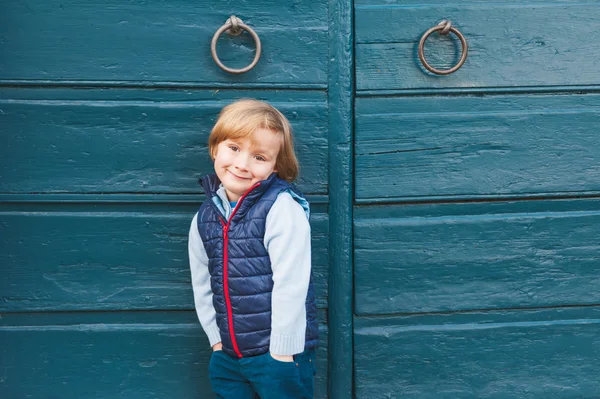 Außenporträt eines niedlichen kleinen Jungen in blauer Weste, der neben einer blauen Holztür steht — Stockfoto