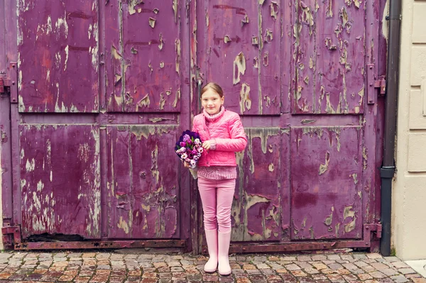 Little girl in a city, wearing bright pink clothes and boots, holding beautiful bouquet of tulips, standing next to old purple wall, toned image — Stock Photo, Image